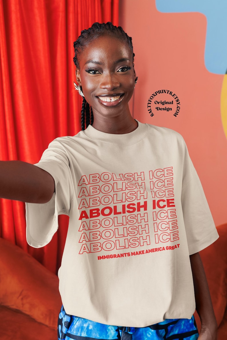 K&ouml;nnte beinhalten: Ein cremefarbenes T-Shirt mit dem Text "ABOLISH ICE" in Rot wiederholt und vertikal gestapelt. Der Satz "IMMIGRANTS MAKE AMERICA GREAT" ist darunter gedruckt. Das Bild zeigt eine l&auml;chelnde Person.