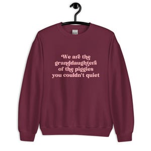 Pode incluir: Um moletom bord&ocirc; com o texto "We are the granddaughters of the piggies you couldn't quiet" em rosa claro, em fonte cursiva. O moletom est&aacute; pendurado em um cabide preto.