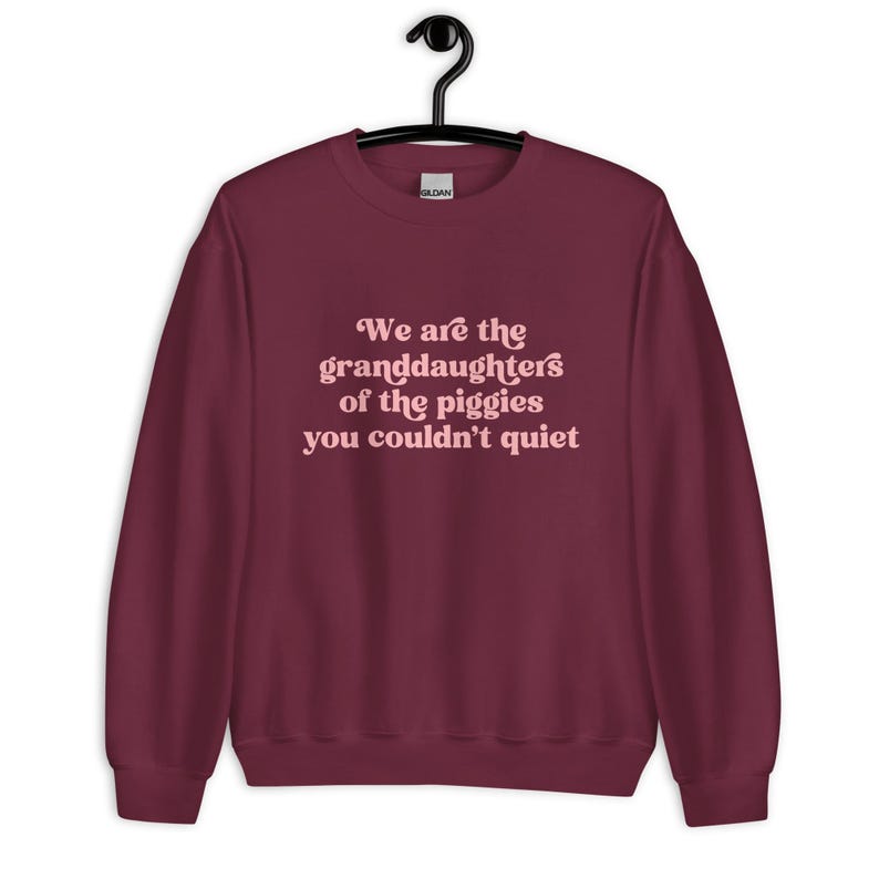 Pode incluir: Um moletom bord&ocirc; com o texto "We are the granddaughters of the piggies you couldn't quiet" em rosa claro, em fonte cursiva. O moletom est&aacute; pendurado em um cabide preto.
