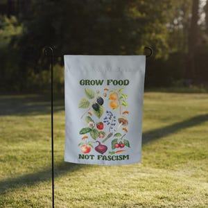 May include: A white garden flag with a green and white illustration of various fruits and vegetables. The text "GROW FOOD NOT FASCISM" is printed in green letters on the flag.