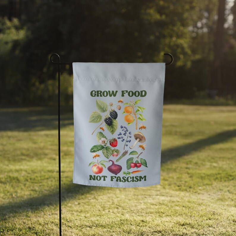 May include: A white garden flag with a green and white illustration of various fruits and vegetables. The text "GROW FOOD NOT FASCISM" is printed in green letters on the flag.