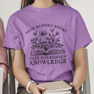 May include: Purple t-shirt with the text "Read Banned Books Gain Forbidden Knowledge" and a black and white illustration of a book with flowers growing out of it.
