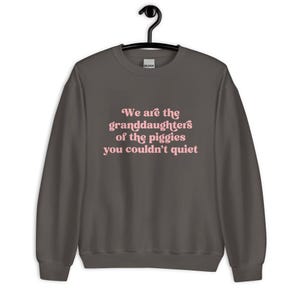 Pode incluir: Camisola cinza escuro com o texto rosa: "We are the granddaughters of the piggies you couldn't quiet". A pe&ccedil;a de roupa est&aacute; pendurada num cabide preto.