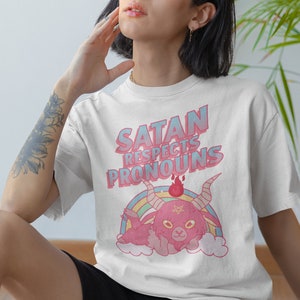 May include: White t-shirt with a pink and blue graphic of a cartoon devil with horns and a pentagram. The text "Satan respects pronouns" is printed in pink and blue letters.