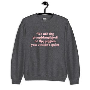 Pode incluir: Camisola cinza escuro com o texto rosa "We are the granddaughters of the piggies you couldn't quiet". A pe&ccedil;a de roupa est&aacute; pendurada num cabide preto.