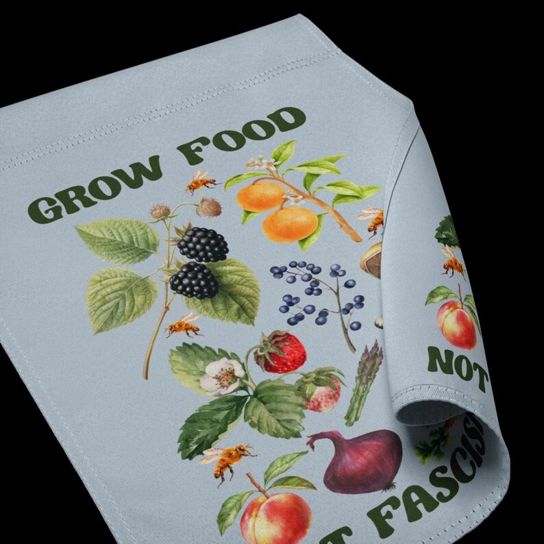 May include: A light blue fabric banner with the text "GROW FOOD NOT FASCISM" in green letters. The banner is decorated with a variety of fruits, vegetables, and flowers, including blackberries, strawberries, peaches, and onions. There are also bees and other insects on the banner.