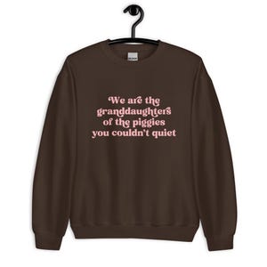 Pode incluir: Um moletom marrom escuro com o texto "We are the granddaughters of the piggies you couldn't quiet" em letras rosa. O moletom est&aacute; pendurado em um cabide preto.