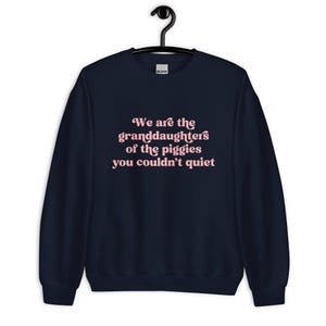Pode incluir: Camisola azul marinho com o texto rosa: "We are the granddaughters of the piggies you couldn't quiet". A pe&ccedil;a de roupa est&aacute; pendurada num cabide preto.