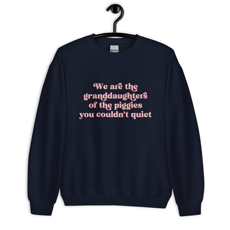 Pode incluir: Camisola azul marinho com o texto rosa: "We are the granddaughters of the piggies you couldn't quiet". A pe&ccedil;a de roupa est&aacute; pendurada num cabide preto.