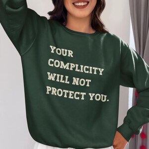May include: A dark green sweatshirt with the text "YOUR COMPLICITY WILL NOT PROTECT YOU." printed in white.