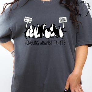 May include: A dark gray t-shirt with a graphic of penguins holding signs that read "I don't even have a job!" and "I'm literally a penguin." The text "PENGUINS AGAINST TARIFFS" is printed below the penguins.