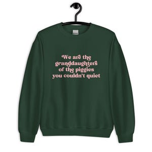 Pode incluir: Uma camisola verde escuro com o texto "We are the granddaughters of the piggies you couldn't quiet" em escrita rosa. A camisola est&aacute; pendurada num cabide preto.