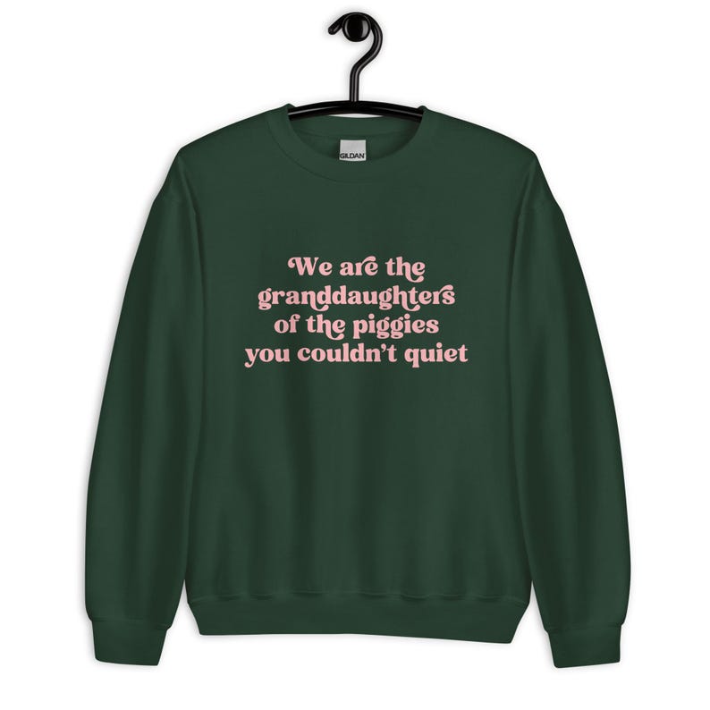 Pode incluir: Uma camisola verde escuro com o texto "We are the granddaughters of the piggies you couldn't quiet" em escrita rosa. A camisola est&aacute; pendurada num cabide preto.