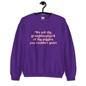 Pode incluir: Um moletom roxo com o texto "We are the granddaughters of the piggies you couldn't quiet" em rosa claro. O moletom est&aacute; pendurado em um cabide preto.