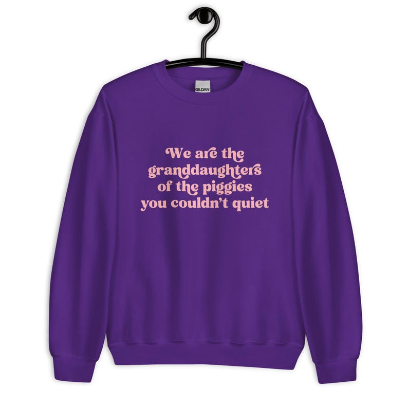 Pode incluir: Um moletom roxo com o texto "We are the granddaughters of the piggies you couldn't quiet" em rosa claro. O moletom est&aacute; pendurado em um cabide preto.