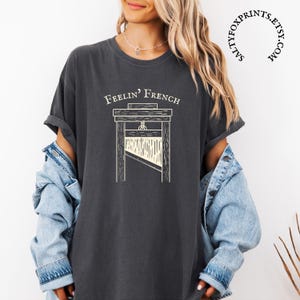 May include: A dark gray t-shirt with a white graphic of a guillotine and the text "Feelin' French".
