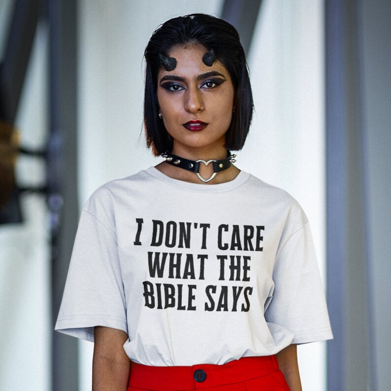 K&ouml;nnte beinhalten: Ein wei&szlig;es T-Shirt mit dem Text "I don't care what the bible says" in schwarzer Schrift. Die Person, die das Shirt tr&auml;gt, hat schwarze Haare und tr&auml;gt eine schwarze Halskette mit einem Herz-Anh&auml;nger.