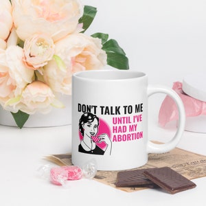 Könnte beinhalten: Eine weiße Keramik-Kaffeetasse mit einer schwarzen und pinkfarbenen Grafik einer Frau, die eine Teetasse hält. Der Text auf der Tasse lautet "Sprich mich nicht an, bevor ich meine Abtreibung hatte."