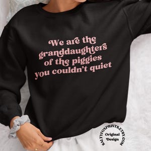 Pode incluir: Camisola preta com a frase rosa "We are the granddaughters of the piggies you couldn't quiet". A camisola &eacute; de gola redonda e mangas compridas. Design de saltyfoxprints.etsy.com.