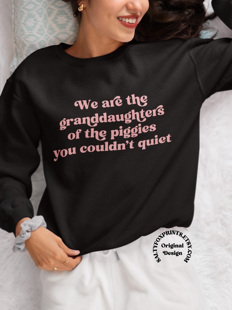 Pode incluir: Camisola preta com a frase rosa "We are the granddaughters of the piggies you couldn't quiet". A camisola &eacute; de gola redonda e mangas compridas. Design de saltyfoxprints.etsy.com.