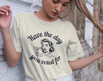 May include: A light beige t-shirt with a black and white illustration of a woman holding a martini glass. The text "Have the day you voted for" is printed on the shirt.