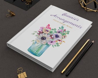 May include: A white journal titled "Bonnie's Arrangements Floral Design Notes" with a watercolor floral design on the cover. The design features a bouquet of flowers in a blue jar. Gold and black pencils and gold binder clips are also visible.