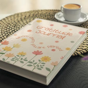 K&ouml;nnte beinhalten: Ein Hardcover-"Gratitude Journal" mit floralem Design und dem Text "bloom where you are planted". Das Journal ist wei&szlig; mit bunten Blumen und liegt auf einem gewebten Platzset. Eine Tasse Kaffee ist im Hintergrund.