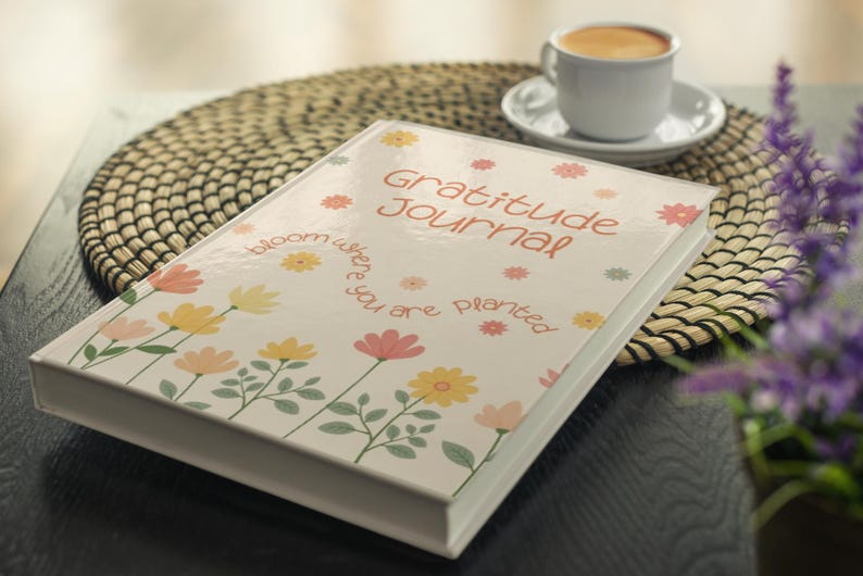 K&ouml;nnte beinhalten: Ein Hardcover-"Gratitude Journal" mit floralem Design und dem Text "bloom where you are planted". Das Journal ist wei&szlig; mit bunten Blumen und liegt auf einem gewebten Platzset. Eine Tasse Kaffee ist im Hintergrund.