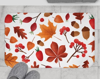 Puede incluir: Una alfombra de baño rectangular con fondo blanco, con un patrón repetido de hojas de otoño, bellotas y bayas en tonos naranja, rojo y marrón. Dos zapatillas grises están en la parte inferior izquierda.