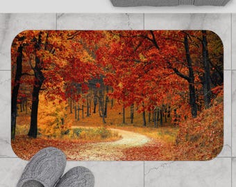 Puede incluir: Alfombra de baño rectangular con una vibrante escena de bosque otoñal. La imagen muestra un camino sinuoso a través de árboles con follaje rojo y naranja. La alfombra está colocada sobre un suelo embaldosado, con un par de zapatillas grises visibles.
