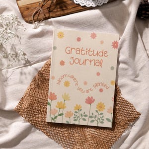 K&ouml;nnte beinhalten: Ein beiges Dankbarkeitstagebuch mit floralen Illustrationen und dem Text "Gratitude Journal". Das Tagebuch liegt auf einem braunen Papiermaterial, mit einem Holztablett, einer Brille, einer Sukkulente und einer Kerze im Hintergrund.