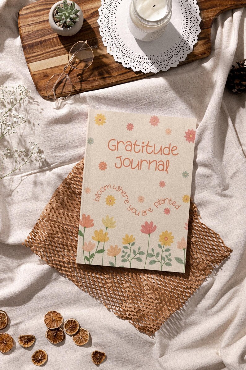 K&ouml;nnte beinhalten: Ein beiges Dankbarkeitstagebuch mit floralen Illustrationen und dem Text "Gratitude Journal". Das Tagebuch liegt auf einem braunen Papiermaterial, mit einem Holztablett, einer Brille, einer Sukkulente und einer Kerze im Hintergrund.
