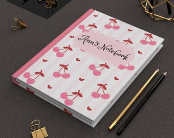 May include: A pink and white striped notebook with a cherry pattern and the text "Ann's Notebook". The notebook has a pink spine and is accompanied by gold and black pencils and gold binder clips.