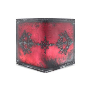 Puede incluir: Cartera roja con acabado envejecido y adornos ornamentales en gris oscuro. La cartera tiene forma rectangular con borde gris oscuro. El diseño presenta un adorno central y detalles en las esquinas.