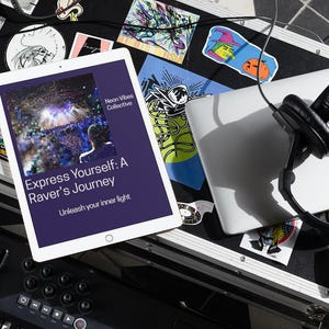 May include: A white tablet displays the book cover "Express Yourself: A Raver's Journey." A laptop, black headphones, and a small keyboard sit on a black surface covered in stickers. The scene suggests music production or DJ equipment.
