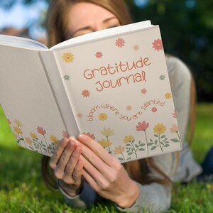 K&ouml;nnte beinhalten: Ein wei&szlig;es Notizbuch mit dem Schriftzug "Gratitude Journal" in Orange, verziert mit floralen Designs. Der Einband zeigt auch den Satz "bloom where you are planted". Das Notizbuch ist ge&ouml;ffnet.