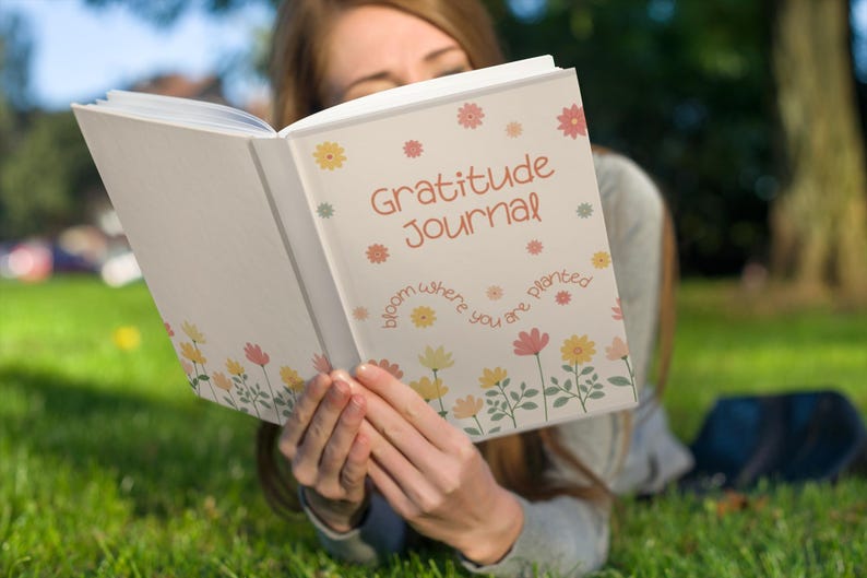 K&ouml;nnte beinhalten: Ein wei&szlig;es Notizbuch mit dem Schriftzug "Gratitude Journal" in Orange, verziert mit floralen Designs. Der Einband zeigt auch den Satz "bloom where you are planted". Das Notizbuch ist ge&ouml;ffnet.