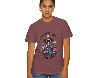 Pode incluir: Uma t-shirt de manga curta cor de malva com um gráfico de um esqueleto com chapéu e jardineiras azuis, segurando uma pá, rodeado por flores cor-de-rosa. O texto "I GARDEN SO I DON'T CHOKE PEOPLE" está impresso a preto à volta do gráfico.