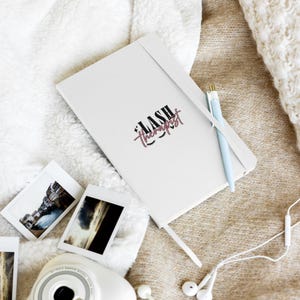May include: A white notebook featuring the black and pink text LASH THERAPIST with small lash icons and star accents. A light blue pen with a gold clip is tucked into the elastic band of the notebook, which is placed alongside two small photos.