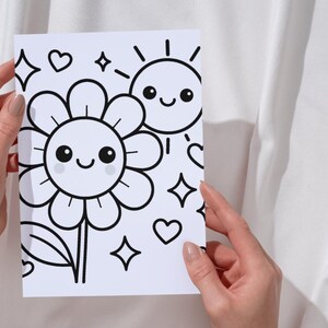Könnte beinhalten: Schwarz-Weiß-Illustration einer lächelnden Blume und Sonne mit Herzen und Sternen. Das Bild wird von zwei Händen vor einem weißen Hintergrund gehalten. Das Kunstwerk eignet sich zum Ausmalen.