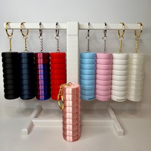 May include: A display of colorful keychains in various colors, including black, navy, red, blue, pink, white, and a translucent design. Each keychain features a cylindrical design with a metal keyring.
