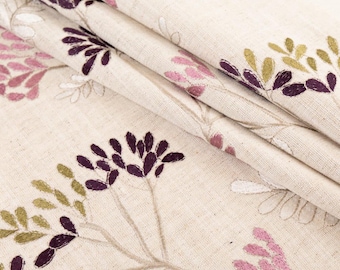 May include: Close-up of beige fabric with embroidered floral design. The embroidery features purple, pink, and green leaves and flowers. The fabric is folded, showing the texture and pattern of the material.