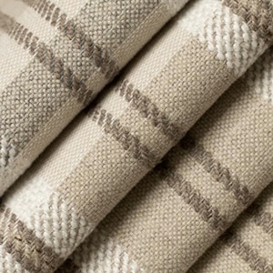 May include: Close-up of folded fabric with a plaid pattern. The fabric features a neutral color palette of beige, cream, and brown, creating a textured appearance. The plaid design is formed by intersecting lines, adding visual interest to the textile.