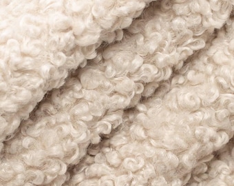 May include: Close-up of a cream-colored, textured fabric with a curly, sheepskin-like appearance. The material is soft and fluffy, with a dense, looped pile. The fabric appears to be folded, creating shadows and depth.