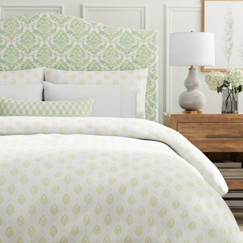 K&ouml;nnte beinhalten: Ein Bett mit einem gr&uuml;n-wei&szlig; gebl&uuml;mten Kopfteil und Bettw&auml;sche. Die Bettw&auml;sche umfasst einen Bettbezug, Kissenbez&uuml;ge und ein Zierkissen. Eine wei&szlig;e Lampe und eine Blumenvase stehen auf einem Nachttisch aus Holz.