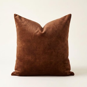 May include: A square throw pillow in a solid, deep chocolate brown color. The fabric has a soft, matte, suede-like texture with a subtle nap. The pillow is plumped to create a slight indentation at the top center, giving it a structured shape.