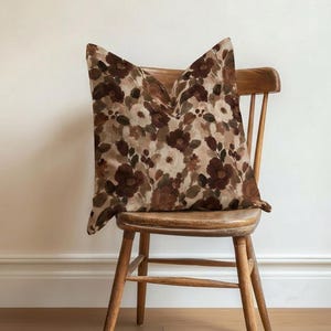 Peut inclure: Un coussin carré orné d'un motif floral dense et pictural dans des tons de brun foncé, moka, beige et crème. Le tissu présente une texture douce et mate avec des motifs de fleurs superposés et des feuilles vert foncé sur l'ensemble.