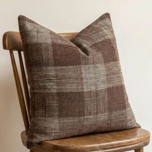 Puede incluir: Un cojín cuadrado con un patrón de cuadros en marrón oscuro y marrón claro con una textura de tela tejida visible. El cojín está colocado sobre una silla de madera con respaldo de listones, mostrando un diseño de cuadrícula en la superficie.