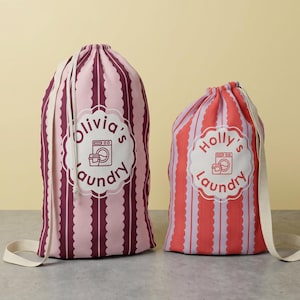 May include: Two drawstring laundry bags. One is pink and burgundy striped with "Olivia's Laundry" and a washing machine graphic. The other is red and pink striped with "Holly's Laundry" and a washing machine graphic. Both have white straps.