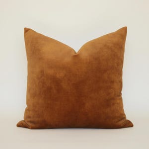 May include: A square throw pillow featuring a solid, rich tobacco brown color. The fabric has a soft, suede-like texture with a matte finish. The pillow is plumped to a firm shape with clean, straight edges and visible seams along the perimeter.